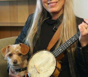 A blonde woman smiles and holds a banjolele. In her lap is a small, yellow dog.