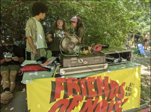 Friends of Noise Bring Music Access to Youth THree young people chat behind a music production console underneath trees on a sunny day