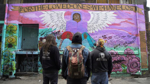 Three people stand in the foreground, backs to camera, looking at a wall mural entitled "For the Loved Ones We Have Lost" that features an angel holding someone in their arms. Three people stand in the foreground, backs to camera, looking at a wall mural entitled "For the Loved Ones We Have Lost" that features an angel holding someone in their arms.