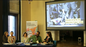 Photo of the panel during Viktoria Kalyniuk's presentation, the slide projected behind is an image of a protester being brutally taken down by heavily armored riot police with the caption "Your State is Protecting You". Photo of the panel during Viktoria Kalyniuk's presentation, the slide projected behind is an image of a protester being brutally taken down by heavily armored riot police with the caption "Your State is Protecting You".