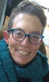 Kate Gray Smiling white woman with short gray hair, blue glasses, and a blue scarf